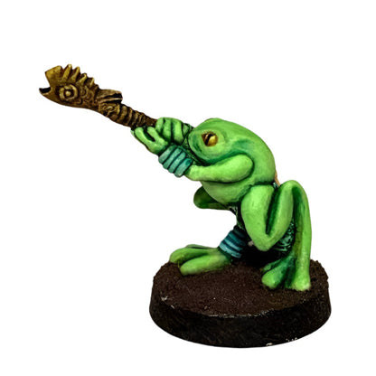 A miniature figurine of a green frog with a blowpipe, designed for tabletop gaming.